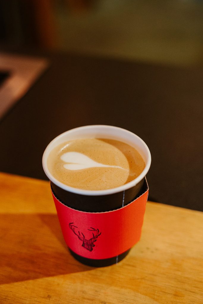 A cup of coffee with heart-shaped latte art and a red sleeve featuring a deer head.